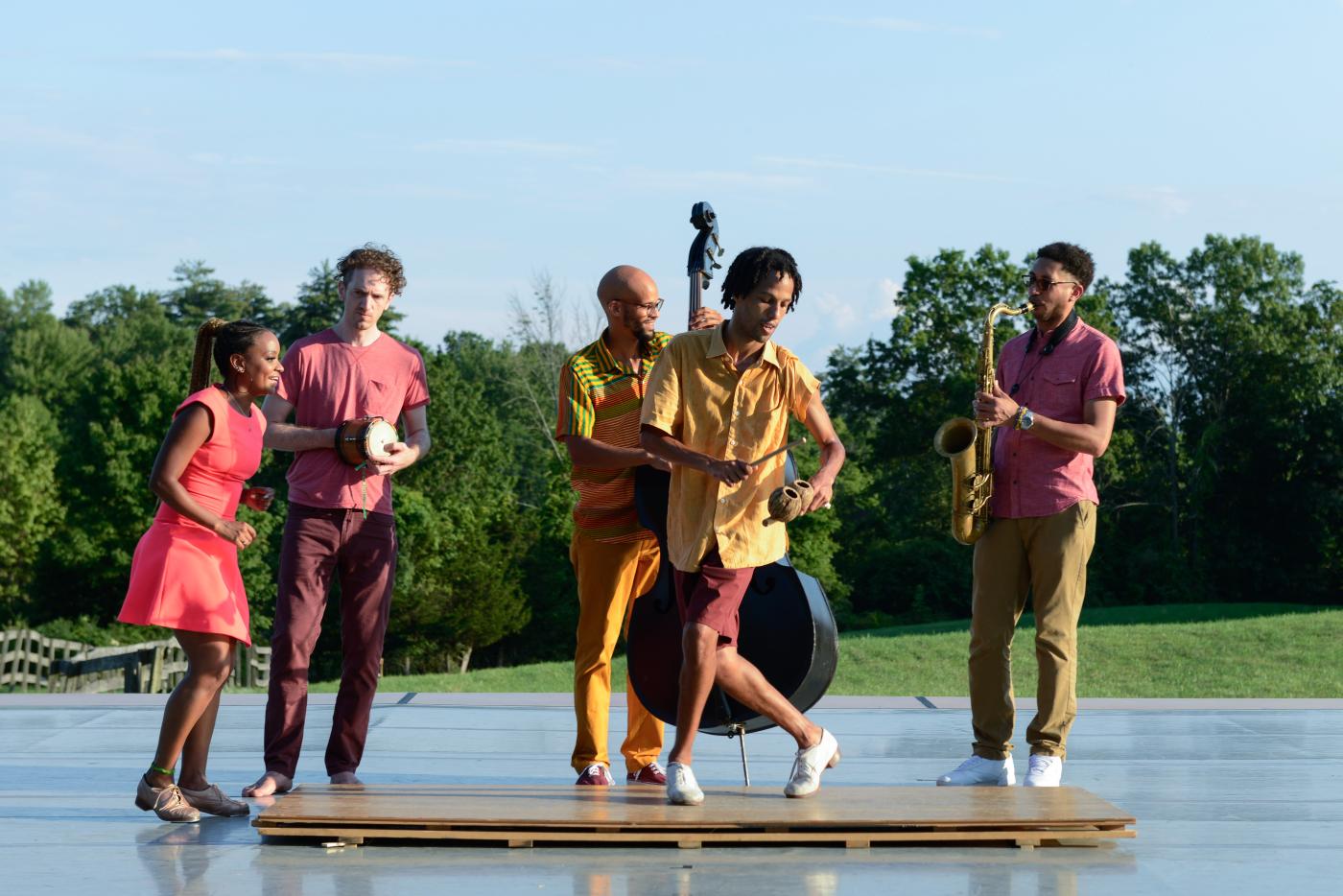 Outside and on a stage, four musicians perform while a dancer steps in front of them.