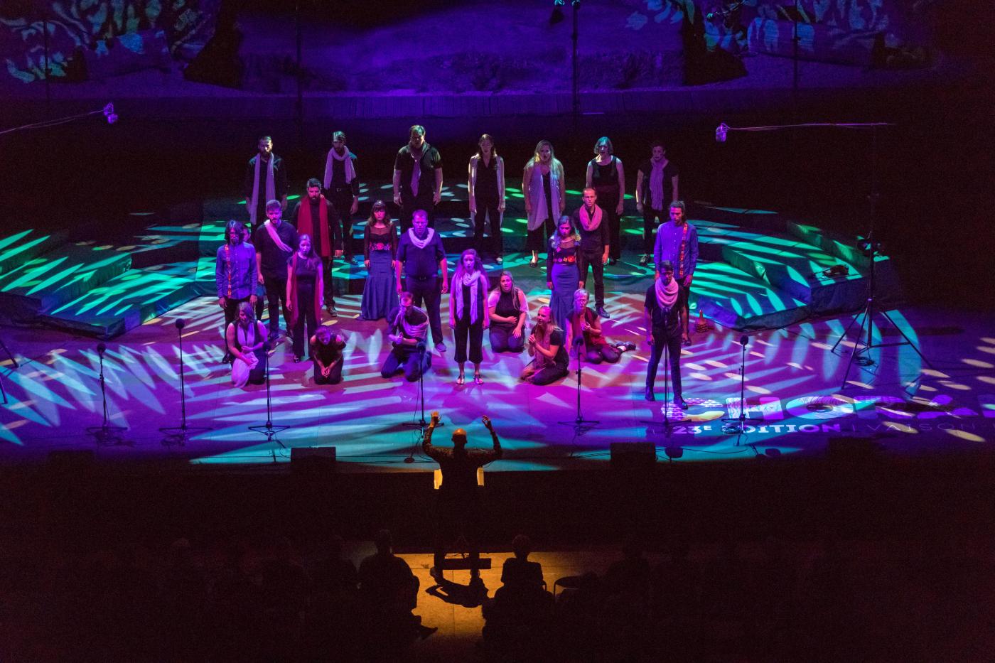 On a spotlight stage, a conductor leads a group of singers in various poses.