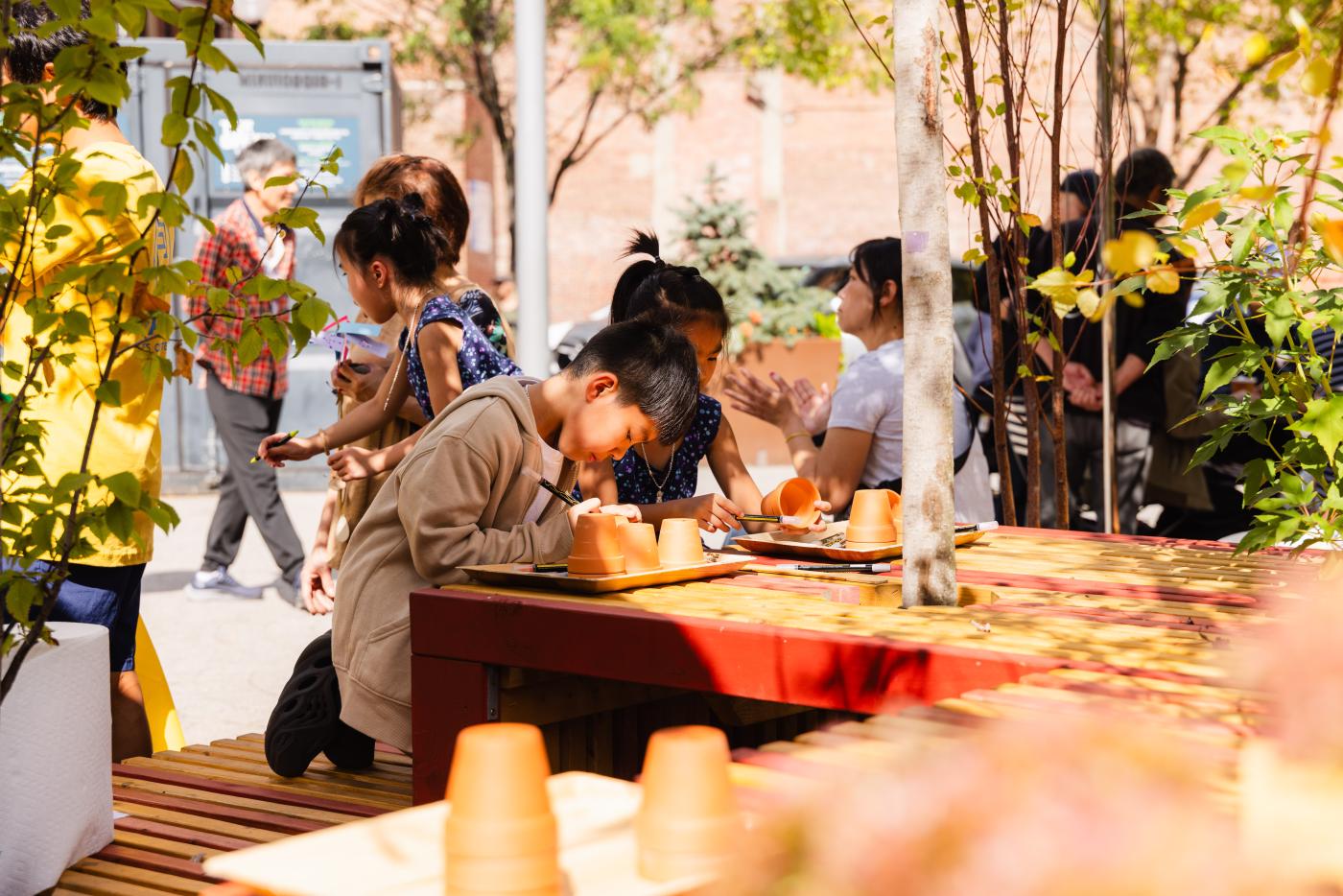 In a park, at a wooden bench with matching tables atop, kids paint clay pots with marker paints.