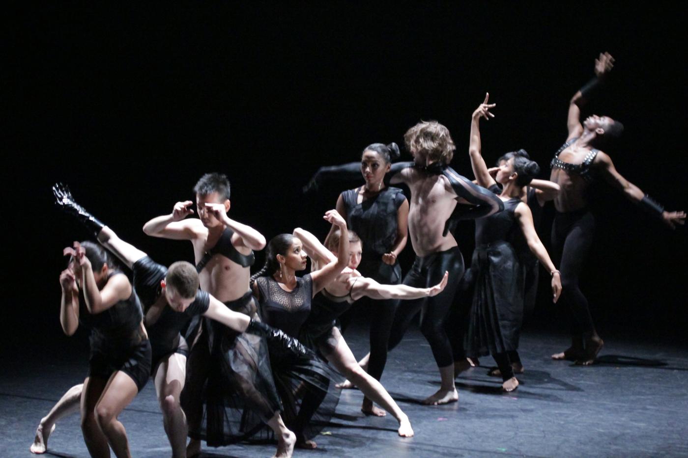 Ten dancers, in front of a black backdrop, perform in a line.