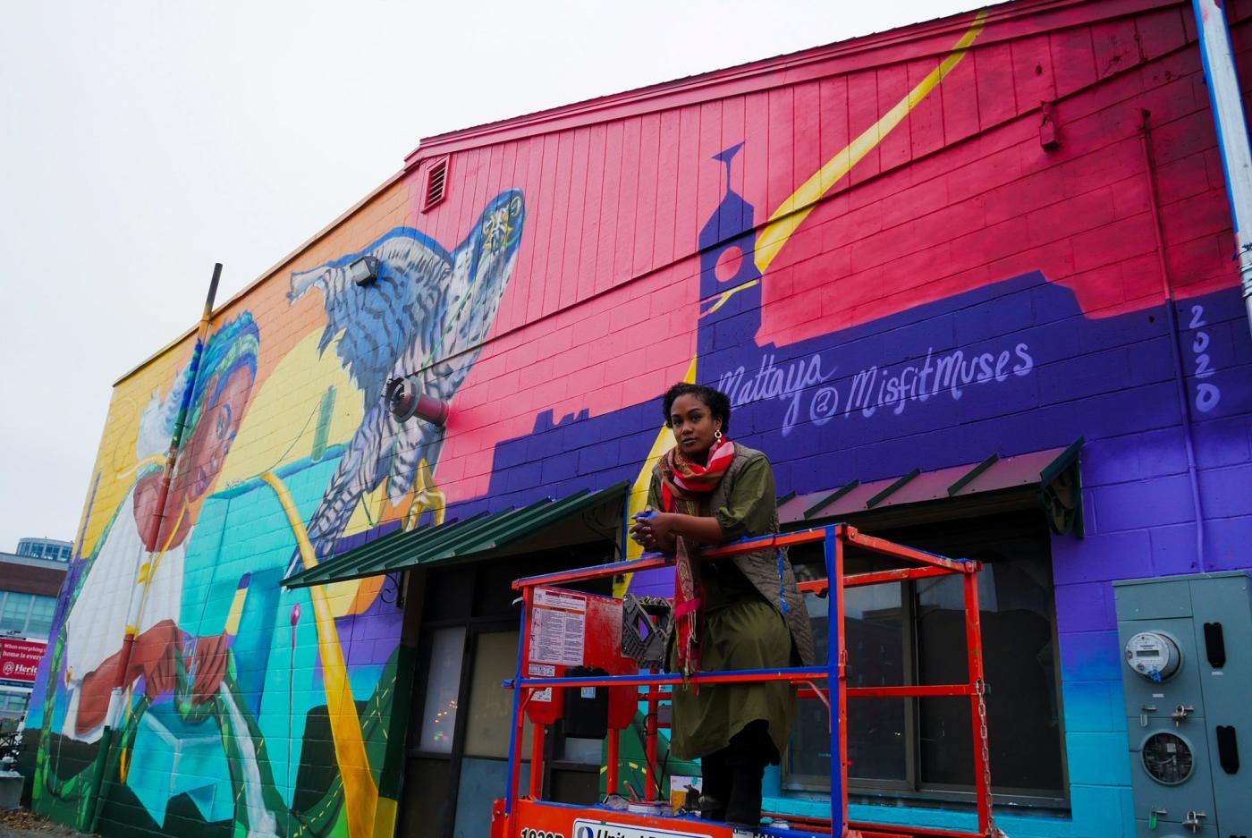 A Black woman poses with a mural behind her, a mural of a Black woman with a bird in very rich, bright colors.