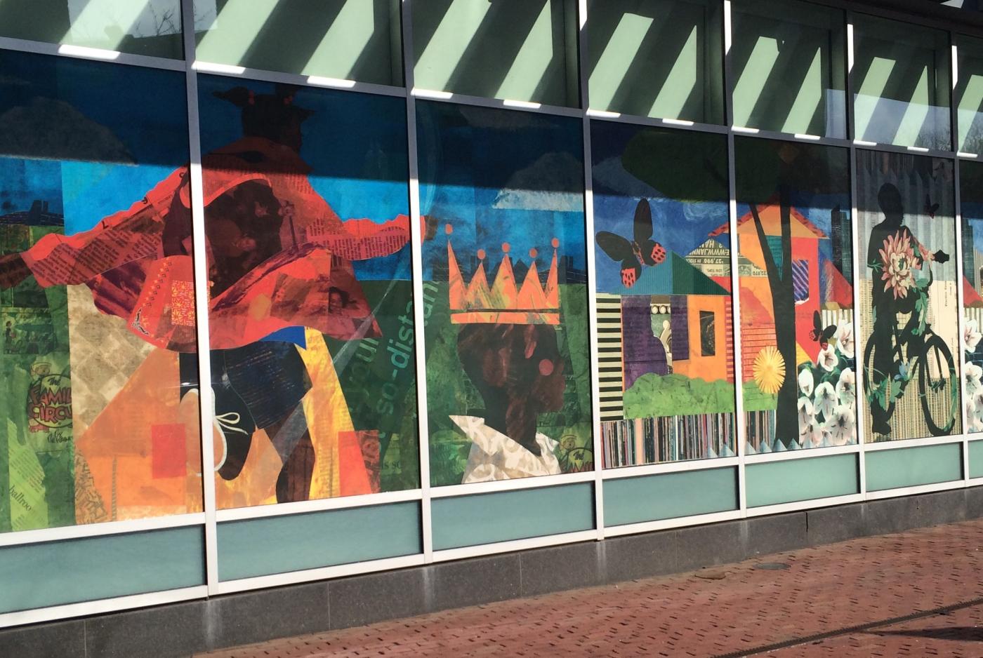 A window mural of Black folks thriving: riding bikes, running through the streets, wearing a crown.