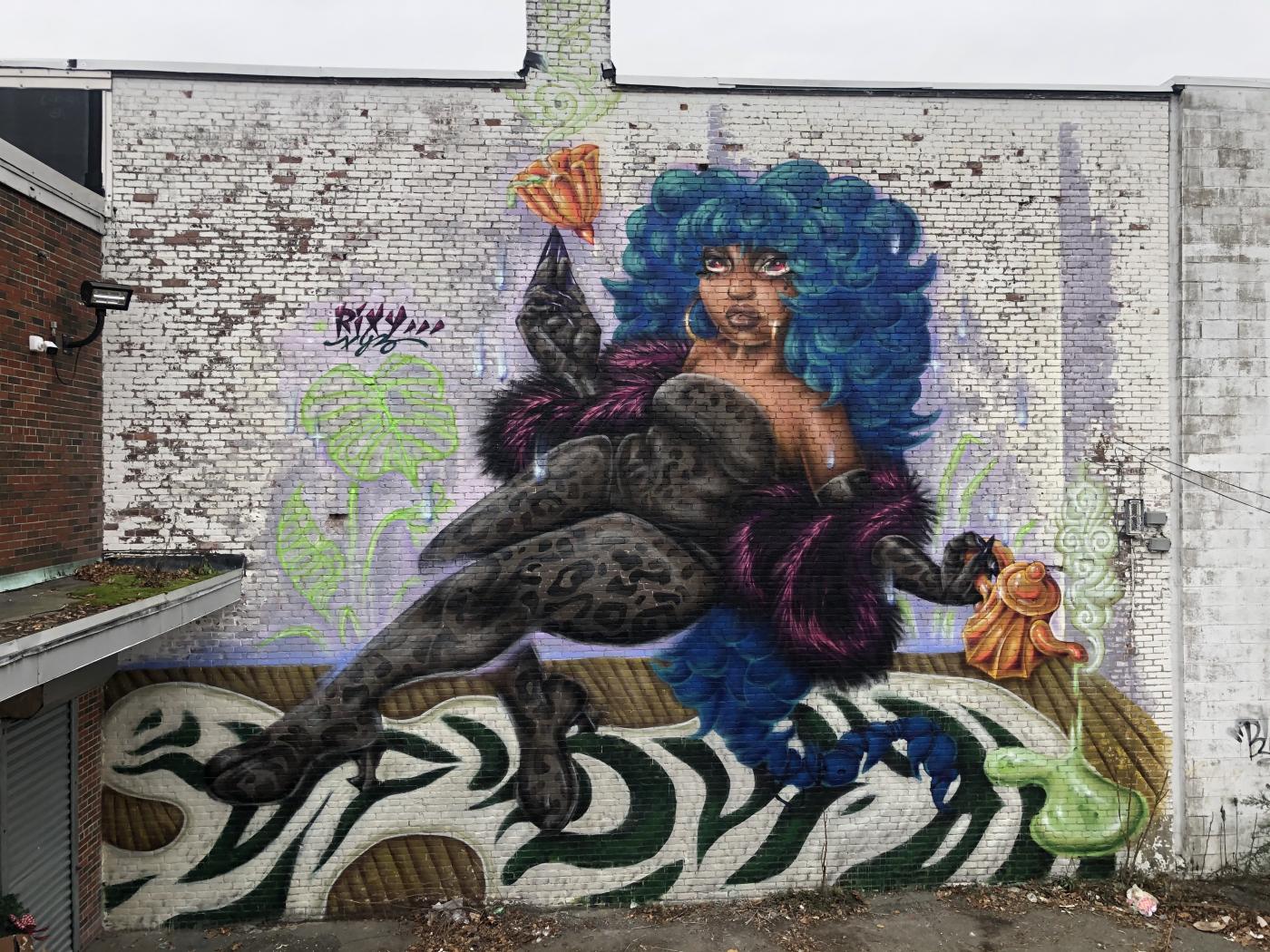 A mural of a woman with blue hair, who wears a leopard print body-suit. She holds a jewel and pours tea.