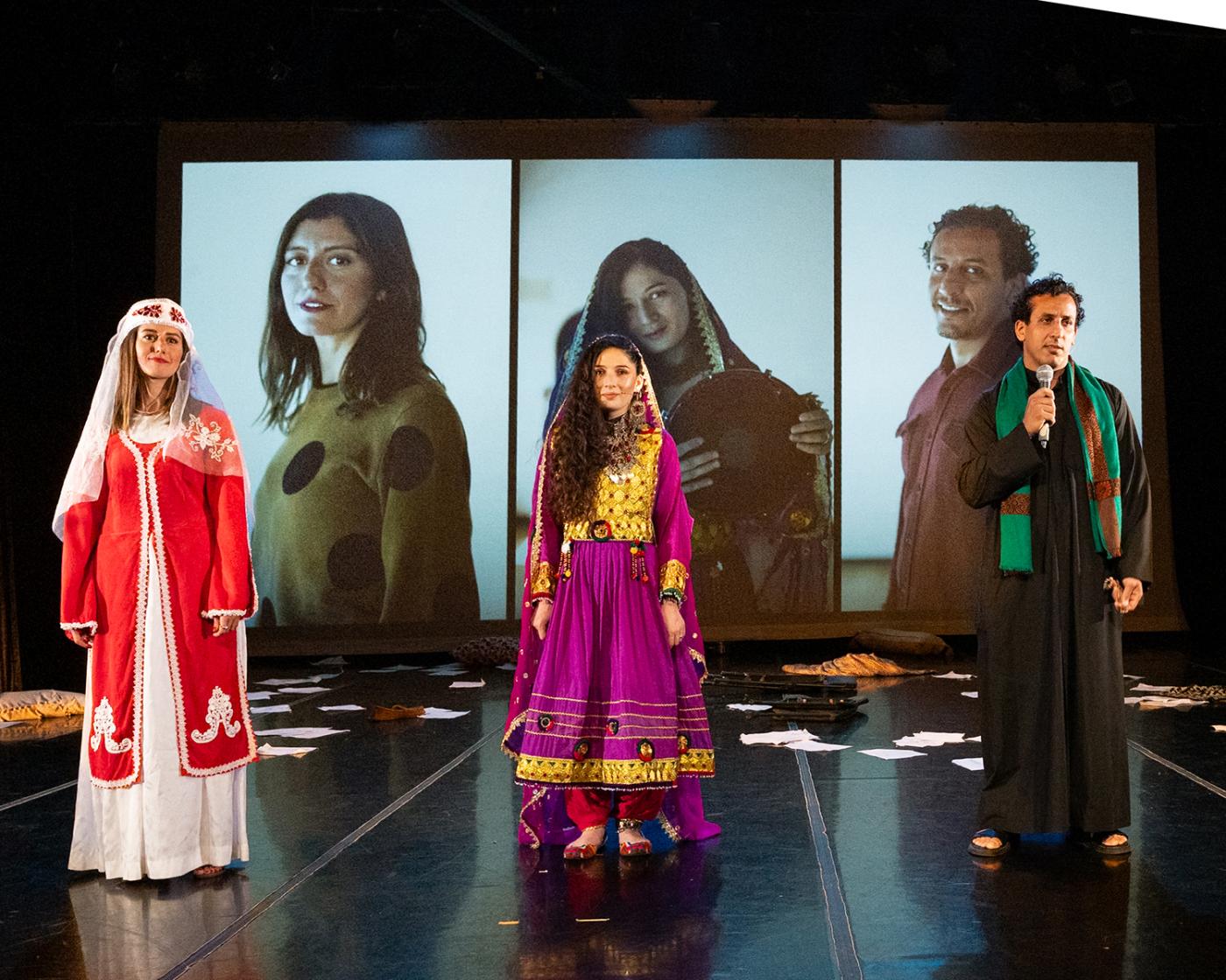 In front of screens with projections of each of the actor's headshots, three performers stand. Two women and a man, who holds a microphone.