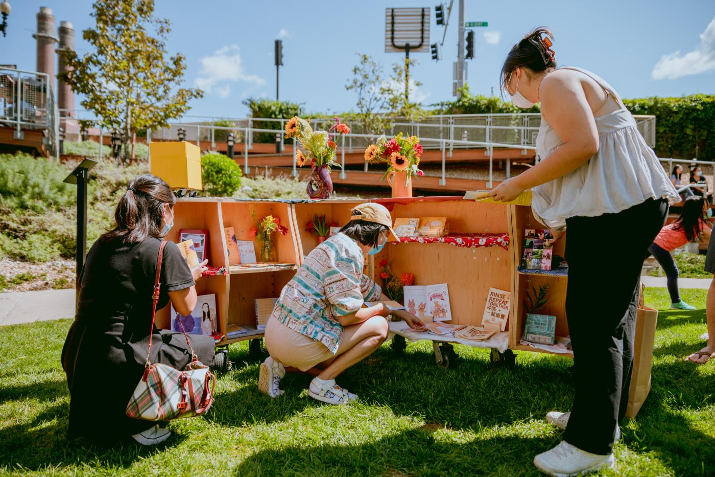 Outside, in a park, three woman poke around an opened box with produce inside it.