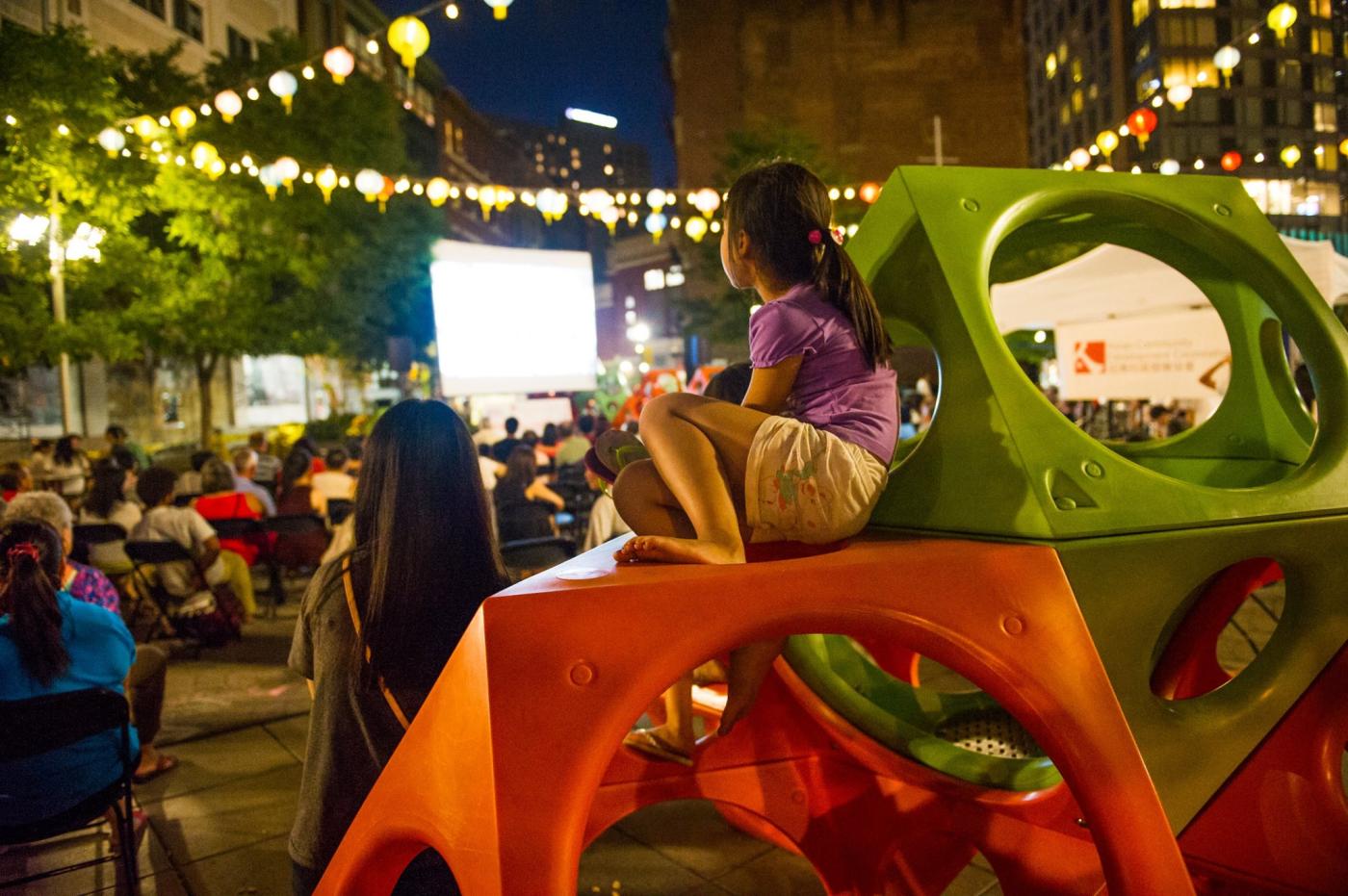 At night, a young Asian girl looks at a projection, while sitting on a jungle gym.