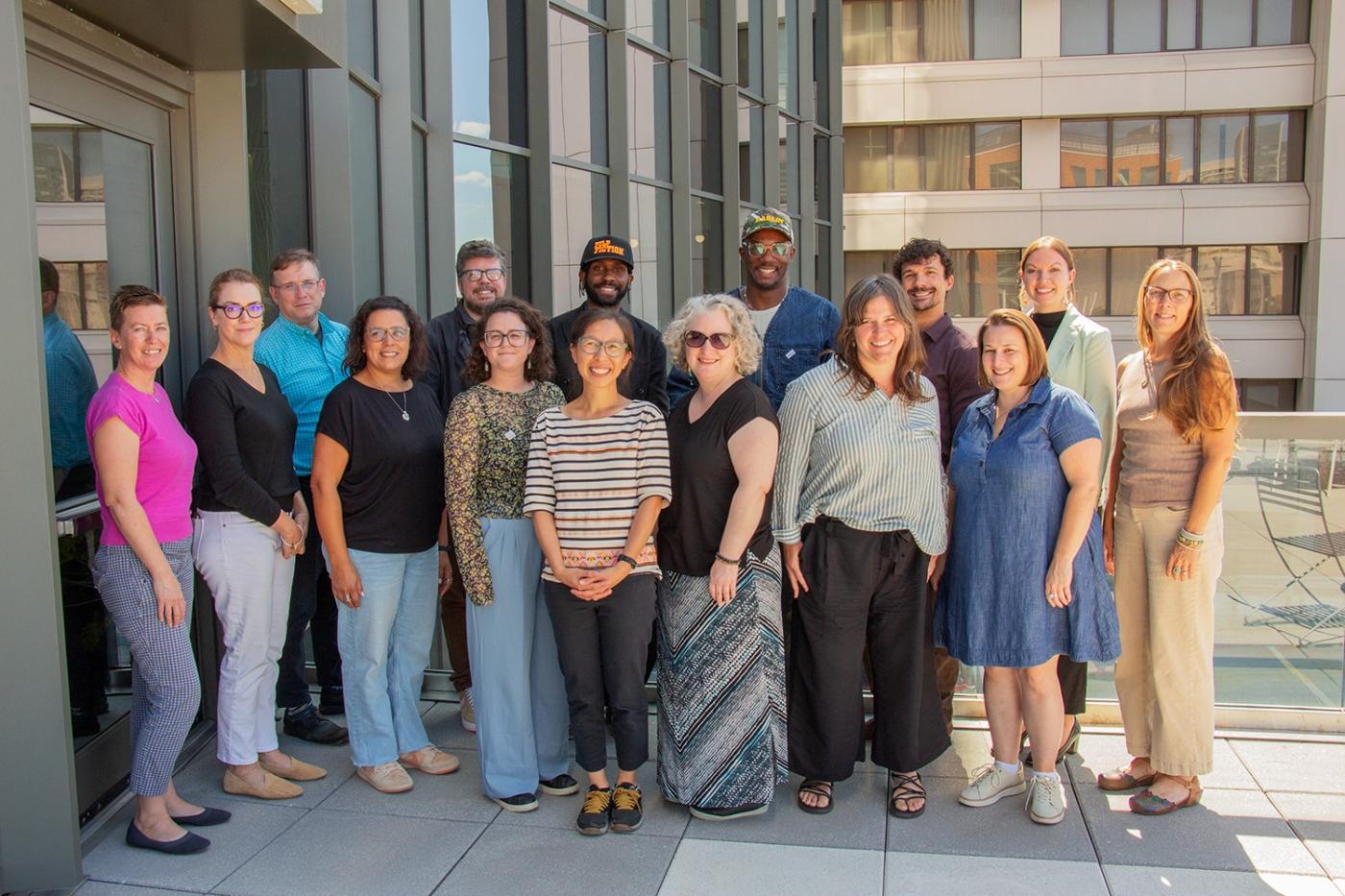 On a concrete deck, fifteen folks pose together as the sun bursts around the corner of the building.
