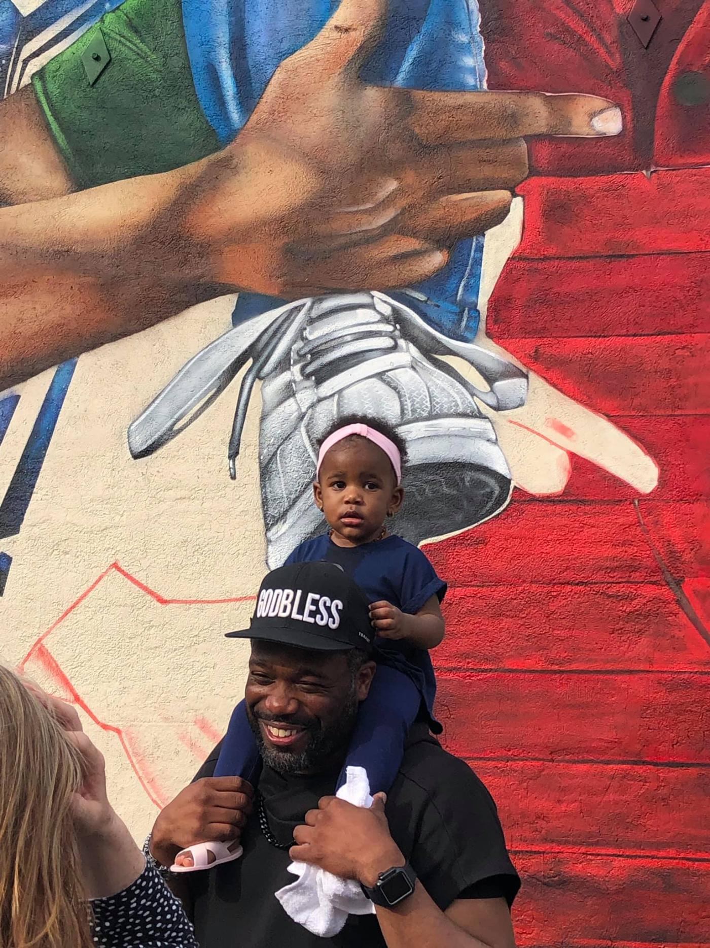 Rob poses with his daughter sitting on his shoulders, in front of one of his murals.