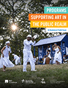 A group of people dressed in white play music and dance on a grassy field. A blue cabin and tall trees form the background, and the sun shines through the trees.