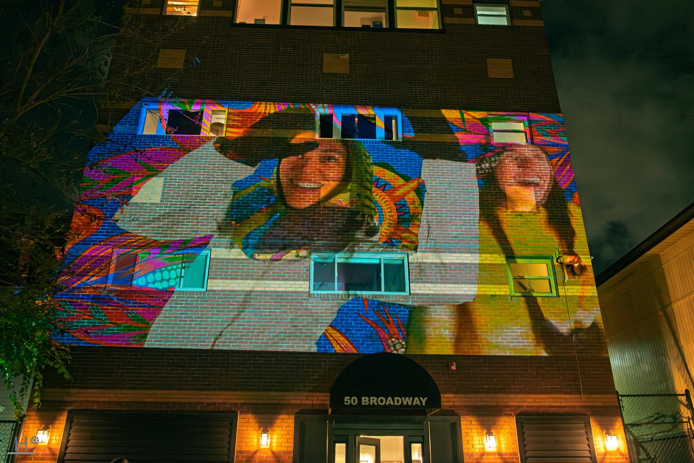 A projection of two women, in winter gloves, on the side of a building at night.