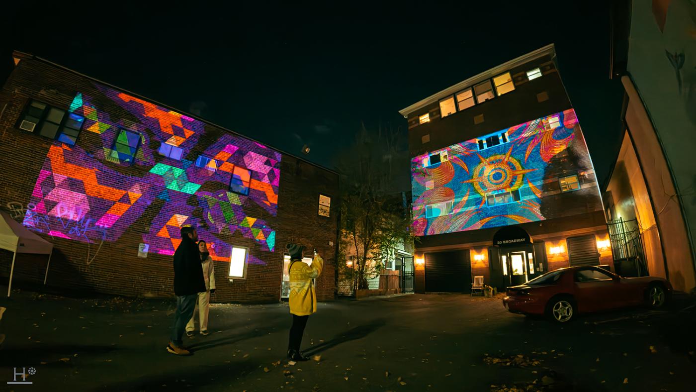 Outside at night, folks take pictures of large scale art projections on the side of buildings.