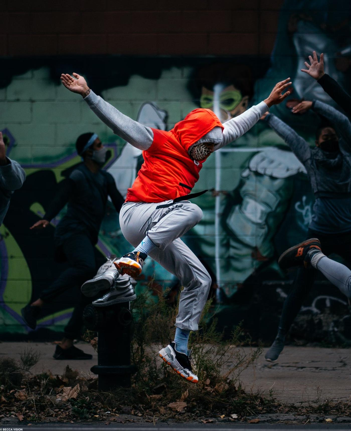 On a street and in front of a mural, a dancer throws their arms and leg up while wearing an orange hoodie.