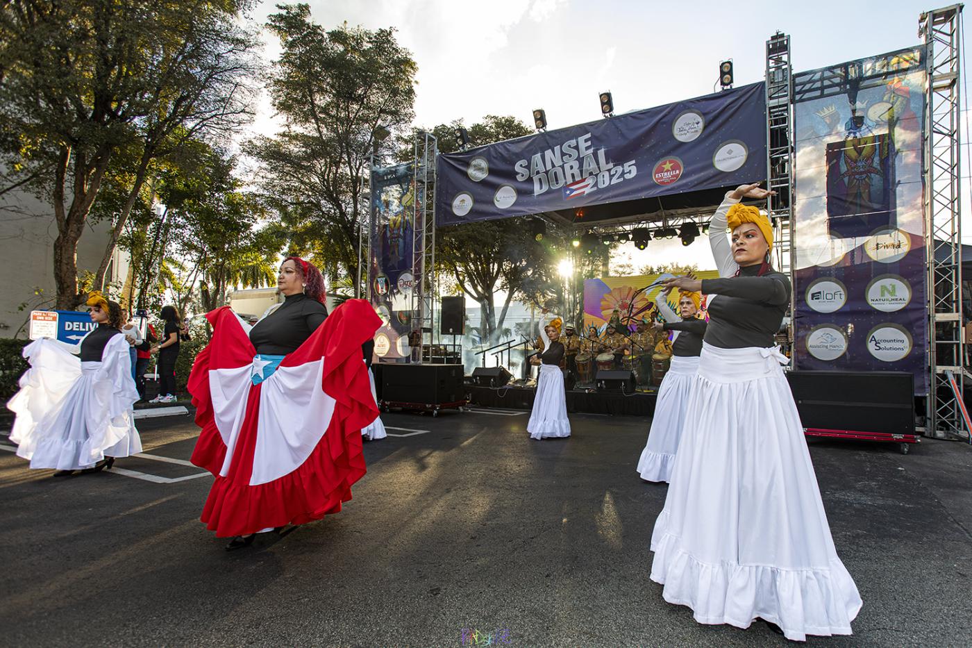 Outdoors and in daylight, four women in long gowns and colorful headwraps dance.