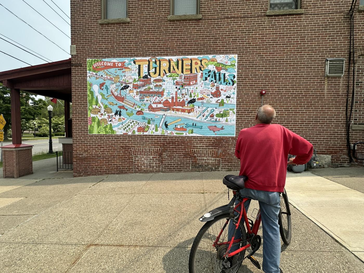 An older man, on a bicycle, admires a wheat paste mural that reads "Turner Falls" and illustrates the town.
