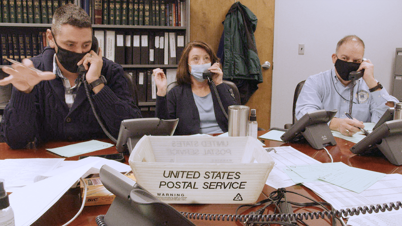 Three people, in masks, sit at a desk and chat on their respective phones.