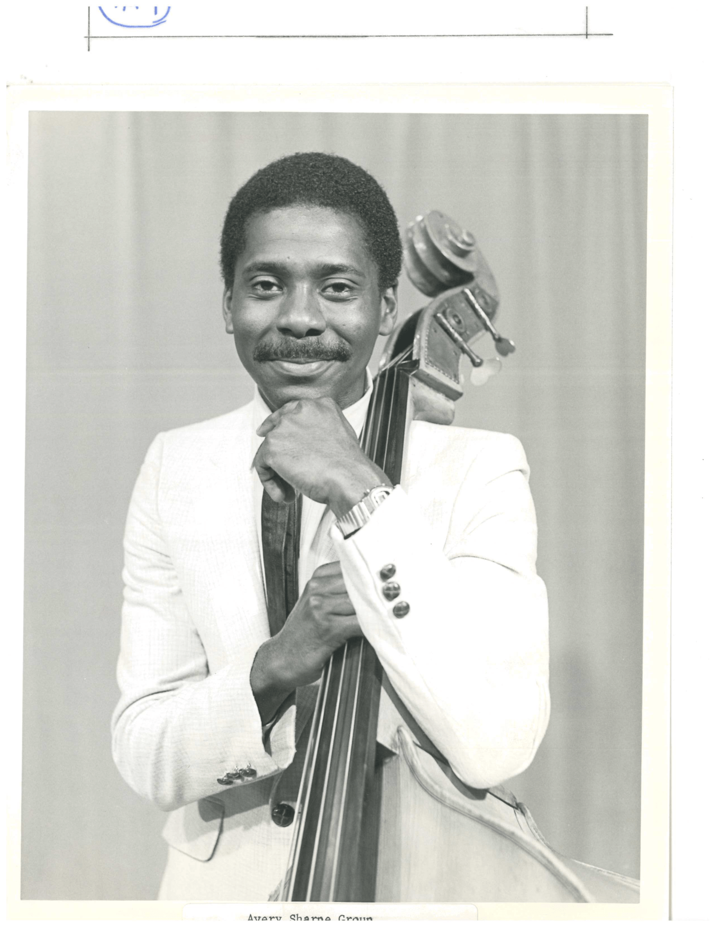 A black and white portrait of a young Avery. He is a Black man in a white suit. He leans on his bass. 