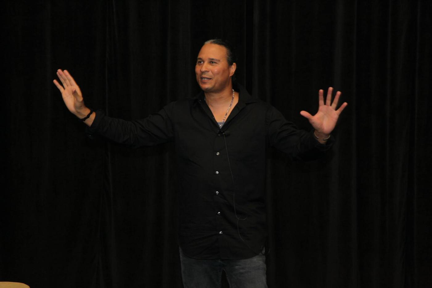 In a black shirt and in front of a black curtain, a man holds his hands up.