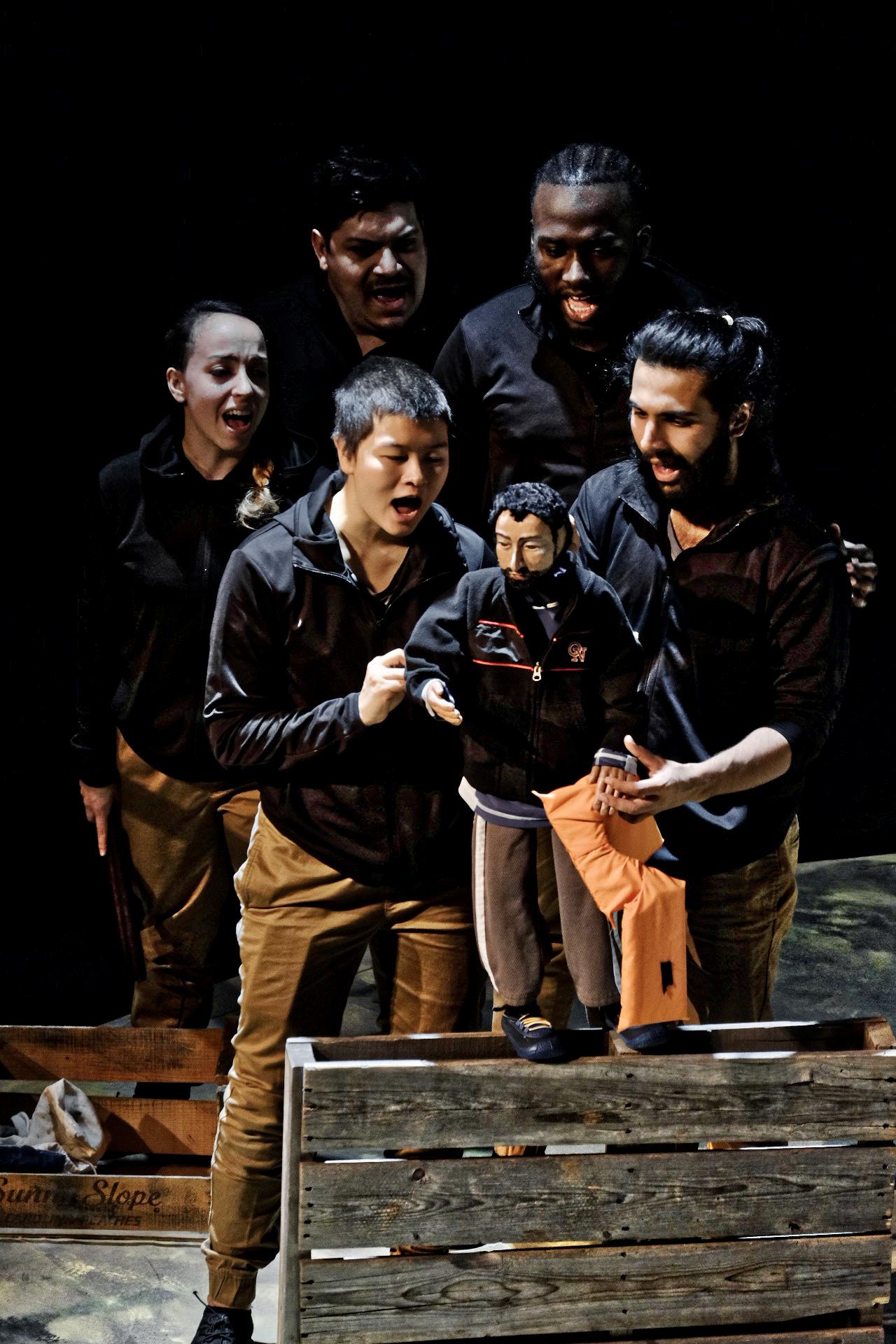 Five performers sing behind a puppet that is handled by two of them.