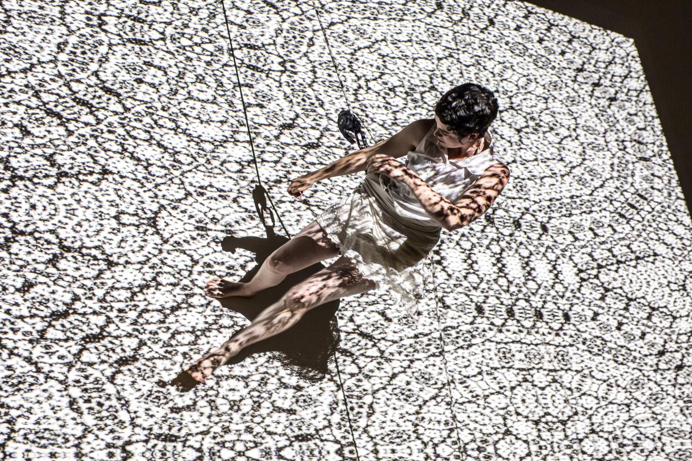 Dangling along a wall with projected patterns over her, a woman dances sideways, on the wall.