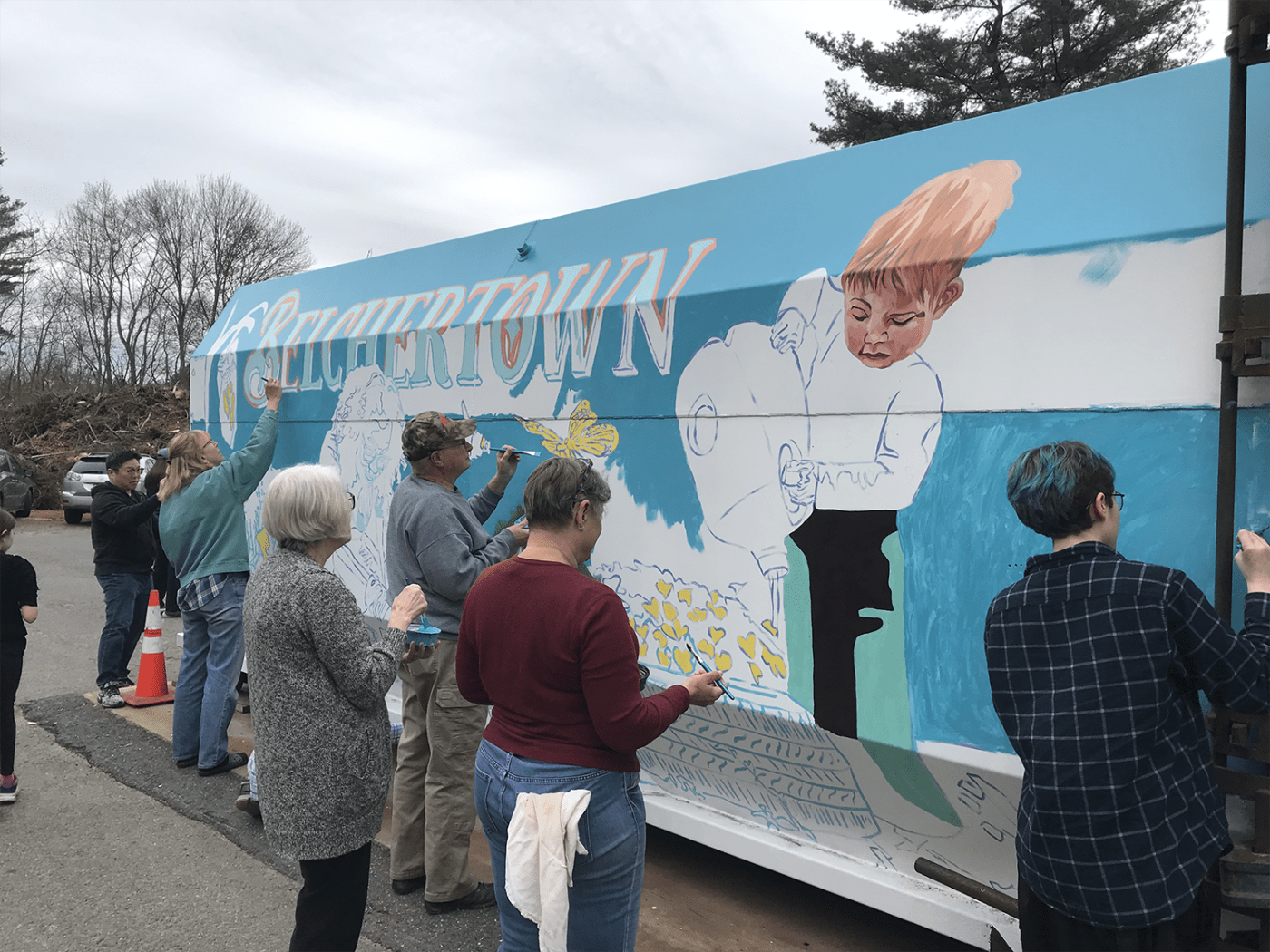 Folks paint the side of a transfer station container. It's cloudy but the right blue of the container pops. In the mural they paint, a young boy pours water over flowers.