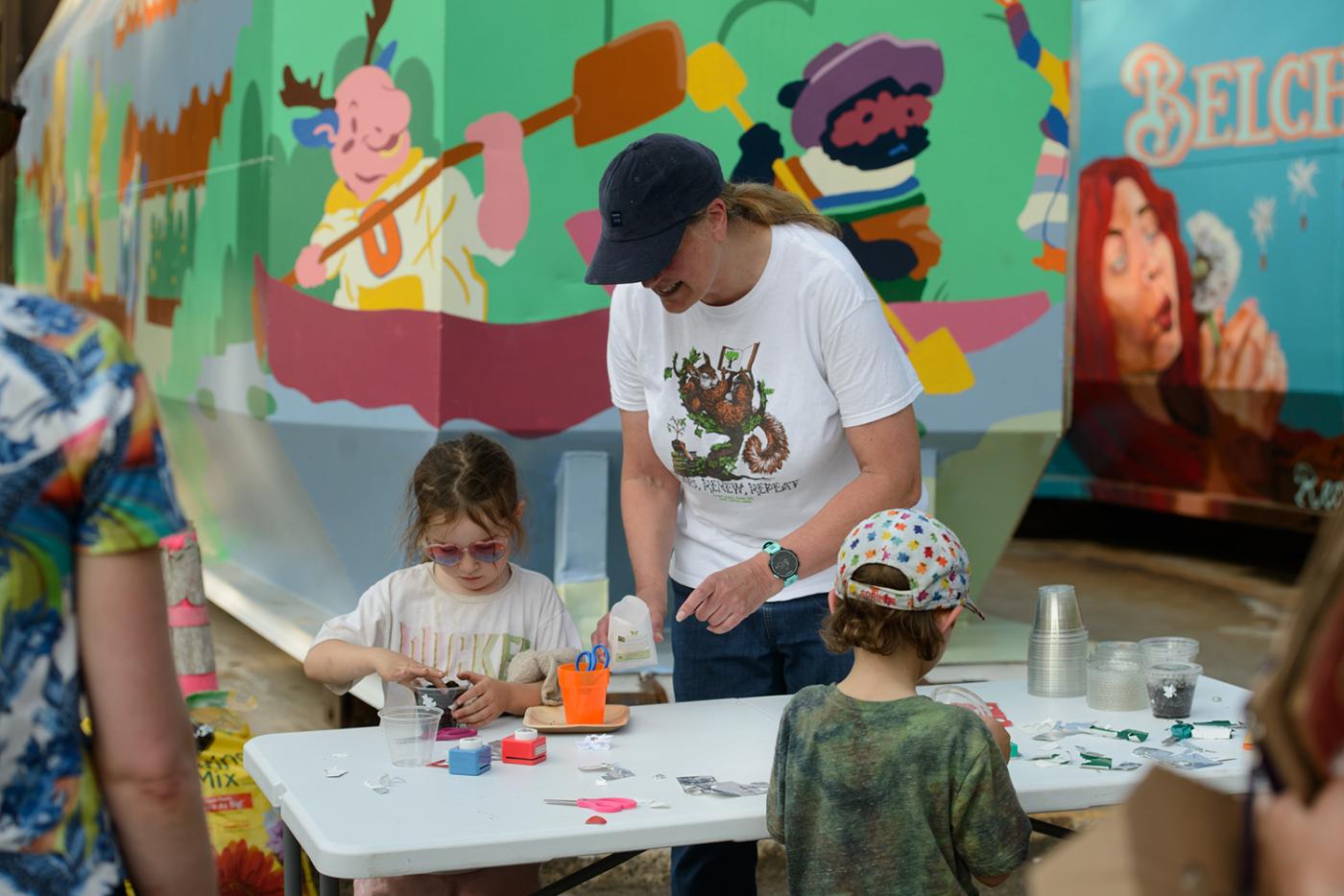 A young woman works with two kids at a craft table. Behind them, more murals on the sides of containers.