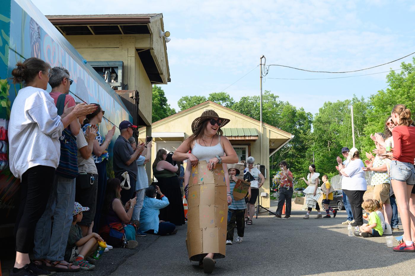 A young woman in a straw hat struts through a crowd. She's fashioned a gown out of cardboard.