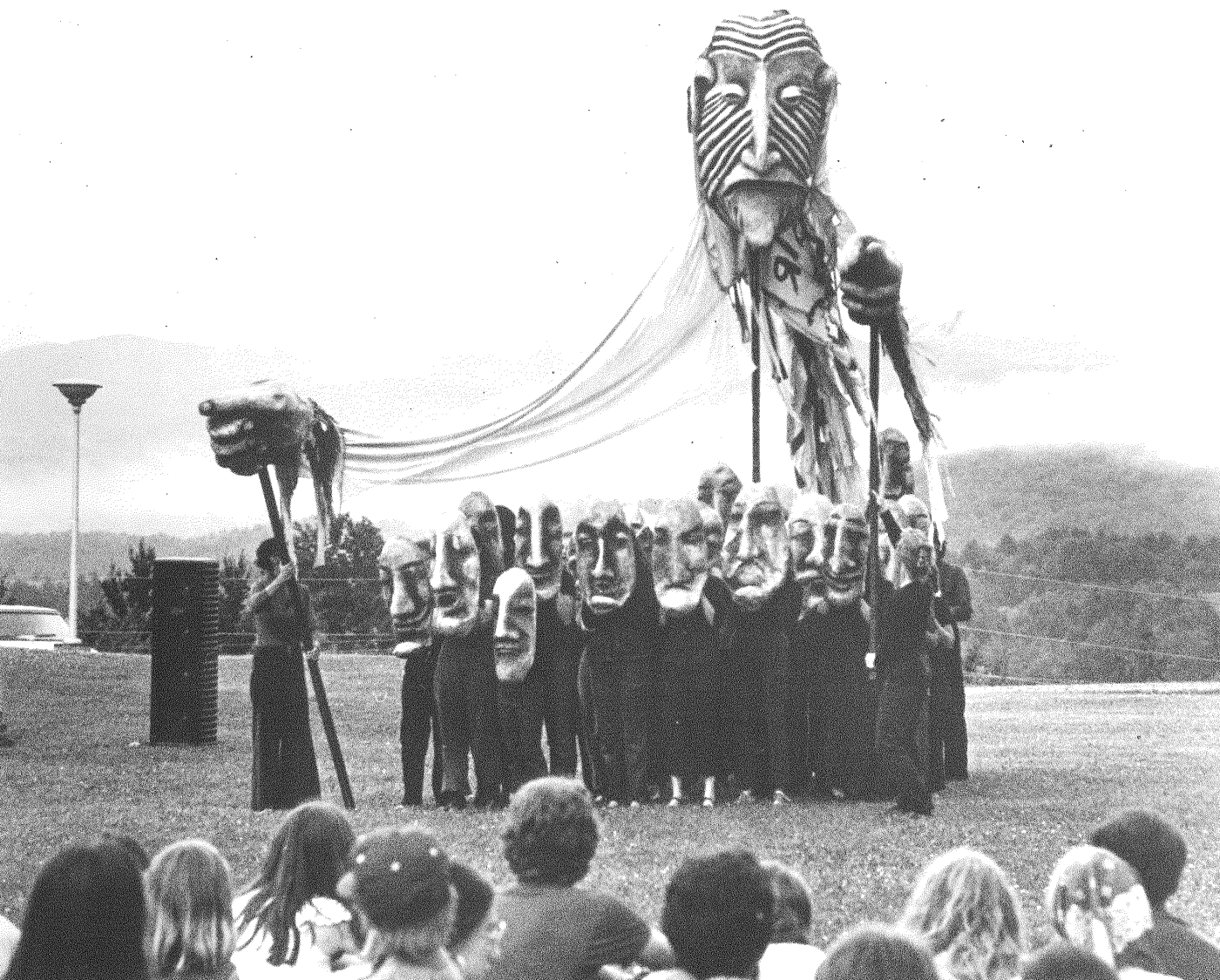 Outdoors, a large group of folks stand with large masks in front of them and another mask with hands connected by strings that marches with them towards an audience.