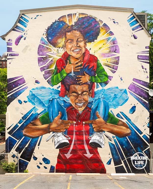 A large colorful mural on the outside of a building featuring a smiling young Black girl sitting on the shoulders of a smiling young Black boy