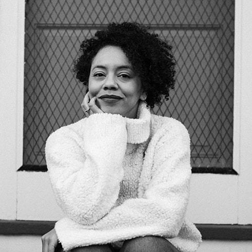 Bulaong has an afro and rests their head on their hand, in a chunky white sweater.