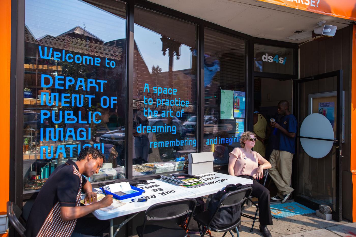 A store front with folks seated at a table. Text on the glass: Welcome to the Department of Public Imagination.