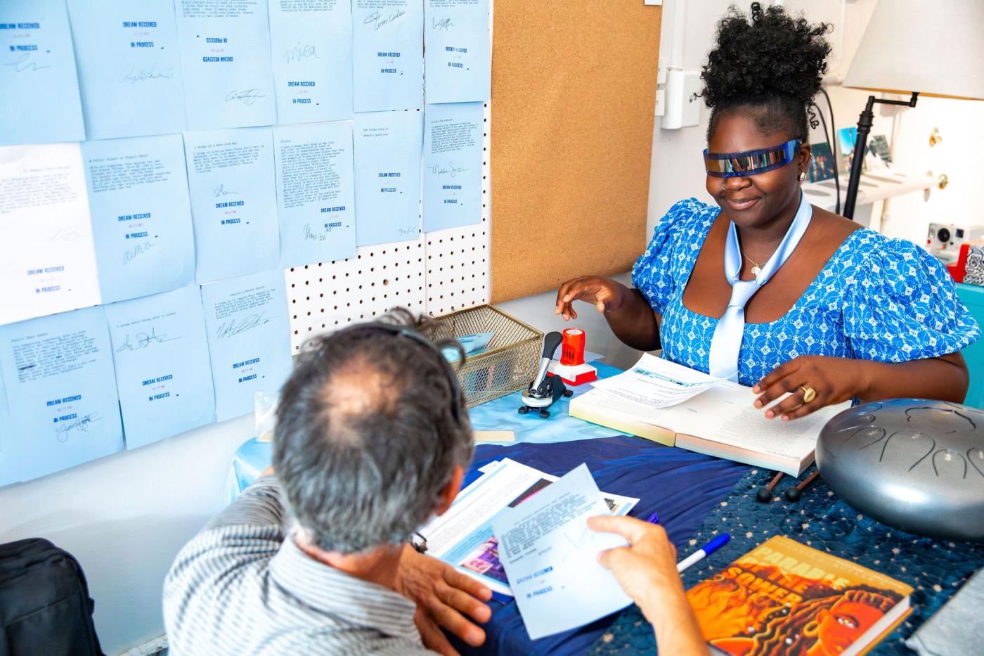 A young woman wears thin glasses over her eyes that may block her vision. She wears a tie around her neck over her dress. Across from her a man with papers in a cramped office space with notes on the wall.