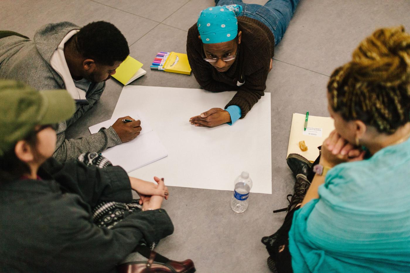 A group of four people sit or lie on the floor while writing on a poster-sized sheet of white paper.