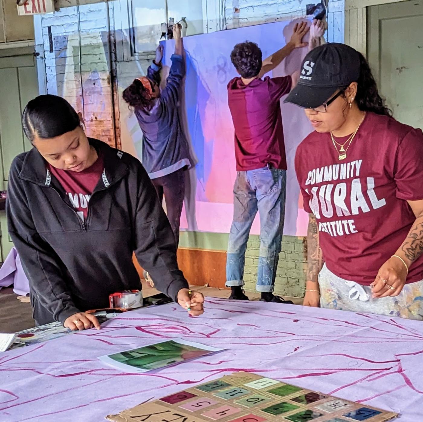 Two young girls of color point to a map on a table, behind them two men hang white paper with art projected onto it.
