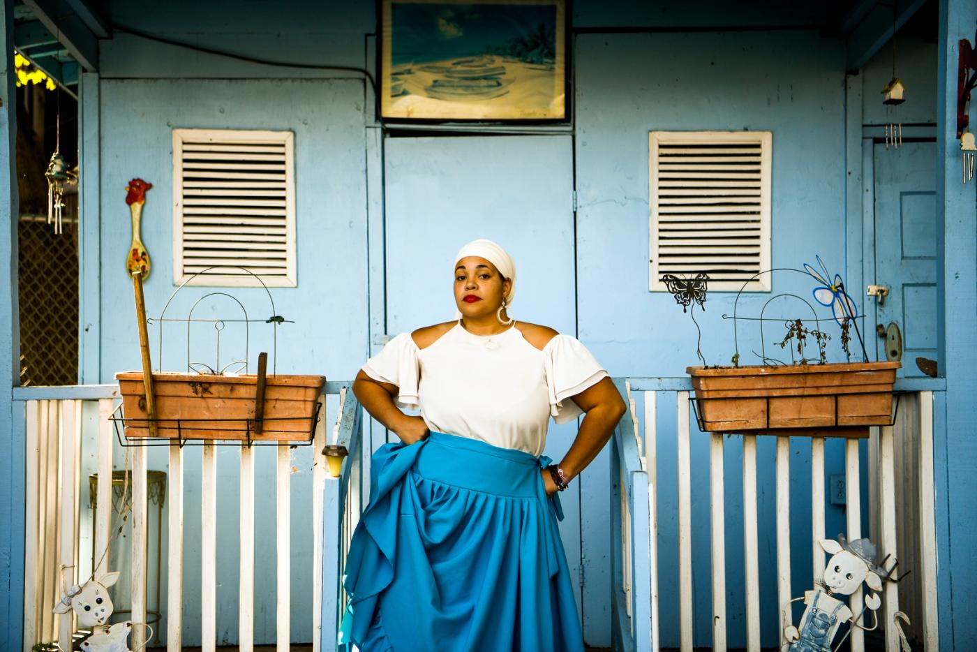 A woman, in a white head wrap and blue dress, stanfds before a doorway with flower boxes and carved lambs.
