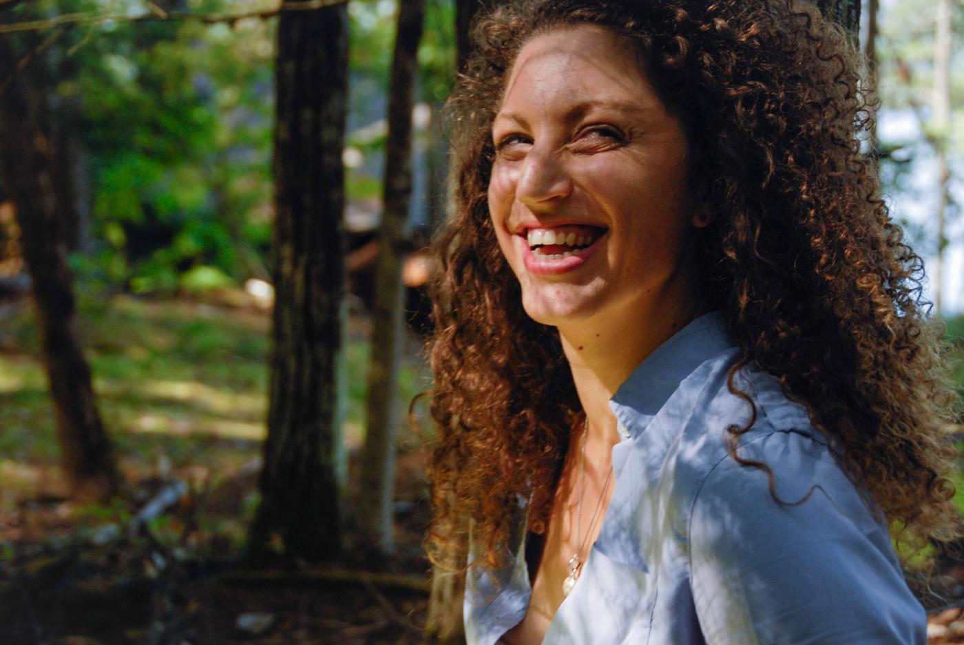 Carla has long curly hair. She smiles and wears a denim shirt, in the woods.