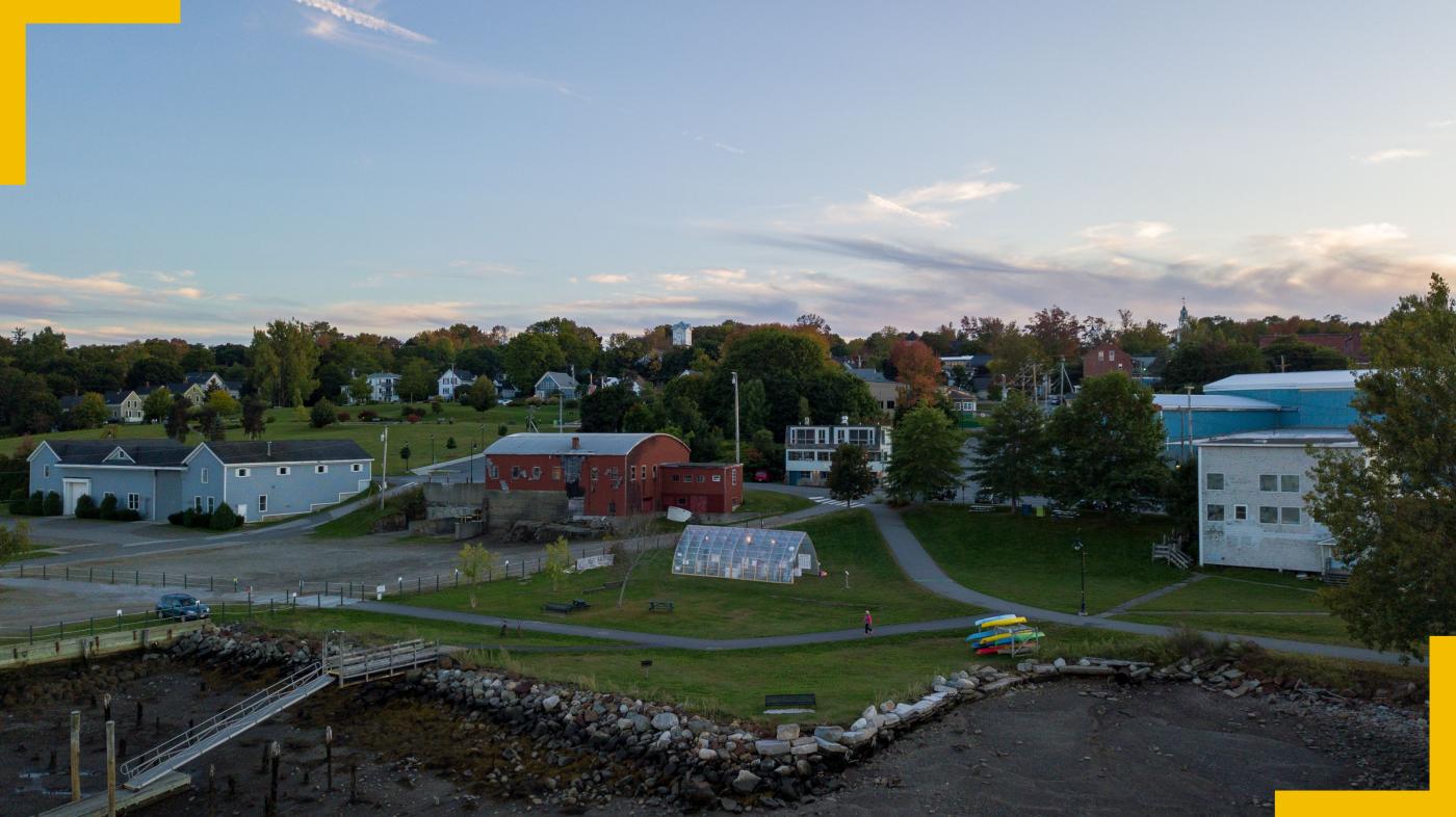 At sunset, along a stony coast lies a barn, some homes, and a clear, wood and plastic boat shed.
