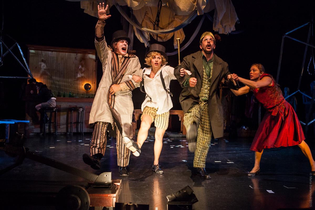 Three people with linked arms on a stage; they are dressed in layers and overcoats and appear to be moving.