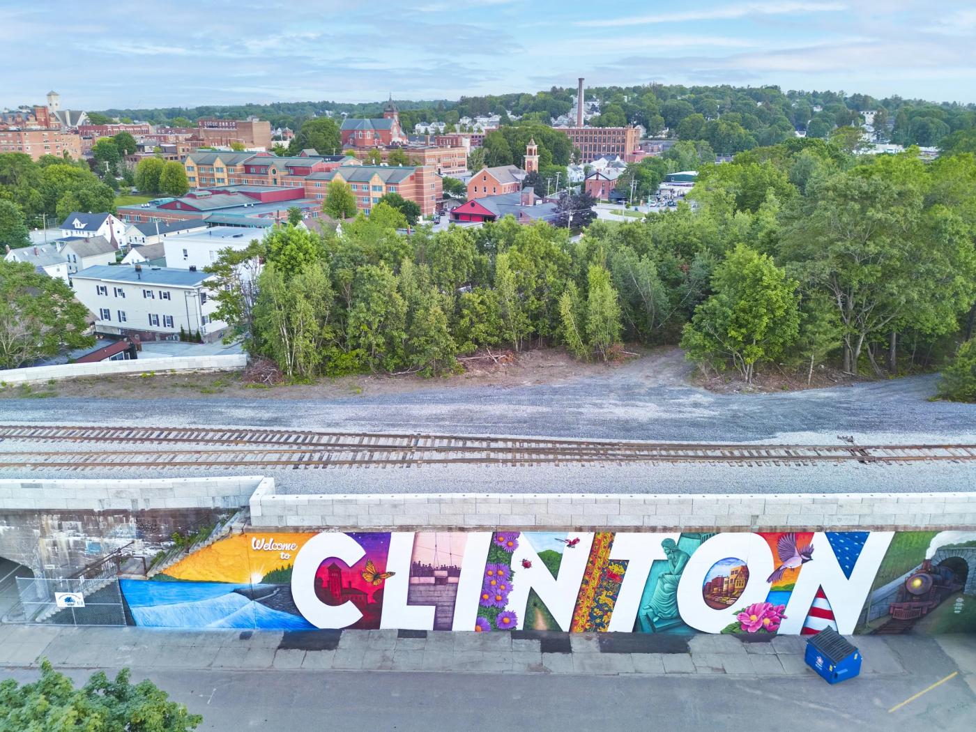 The mural, which spells out Clinton, sits on the side of a wall that creates a bridge for a train. The town of Clinton sits beyond the tracks with tall green trees and a bright blue sky.
