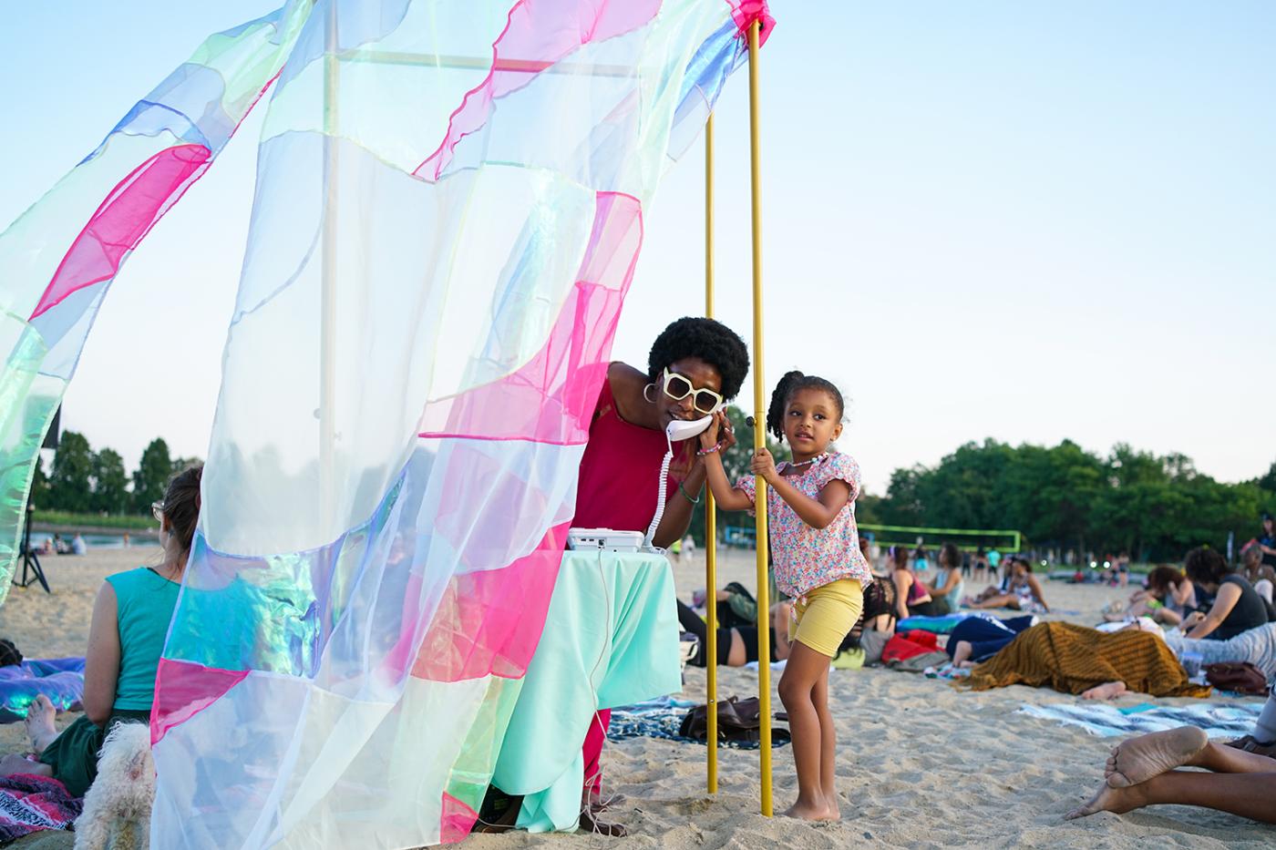 On the beach and within a wall of fabric, a woman holds a phone to her ear. A young girl stands next to her and holds onto the frame of the structure that holds up the wall of colorful fabric.
