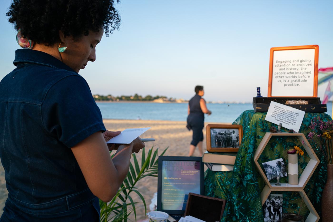 A young woman takes notes while looking at framed text, on the beach. Beyond her, folks walk towards the water.