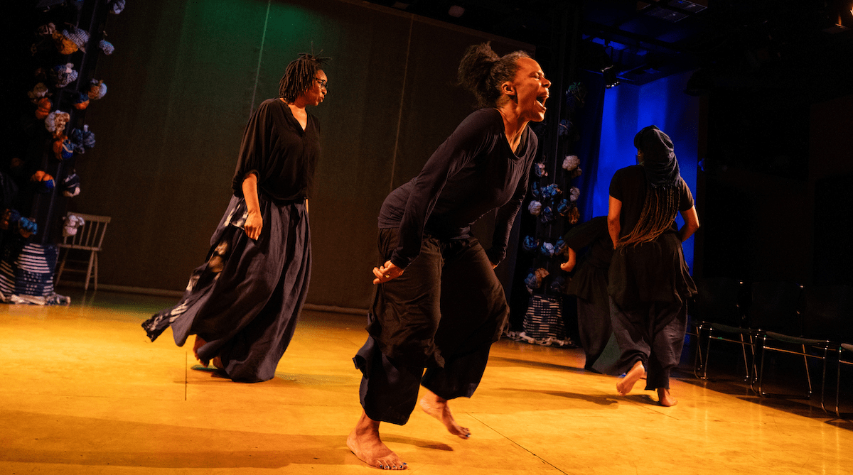 Four performers dressed in black move about a stage. One of them seems to scream passionately.