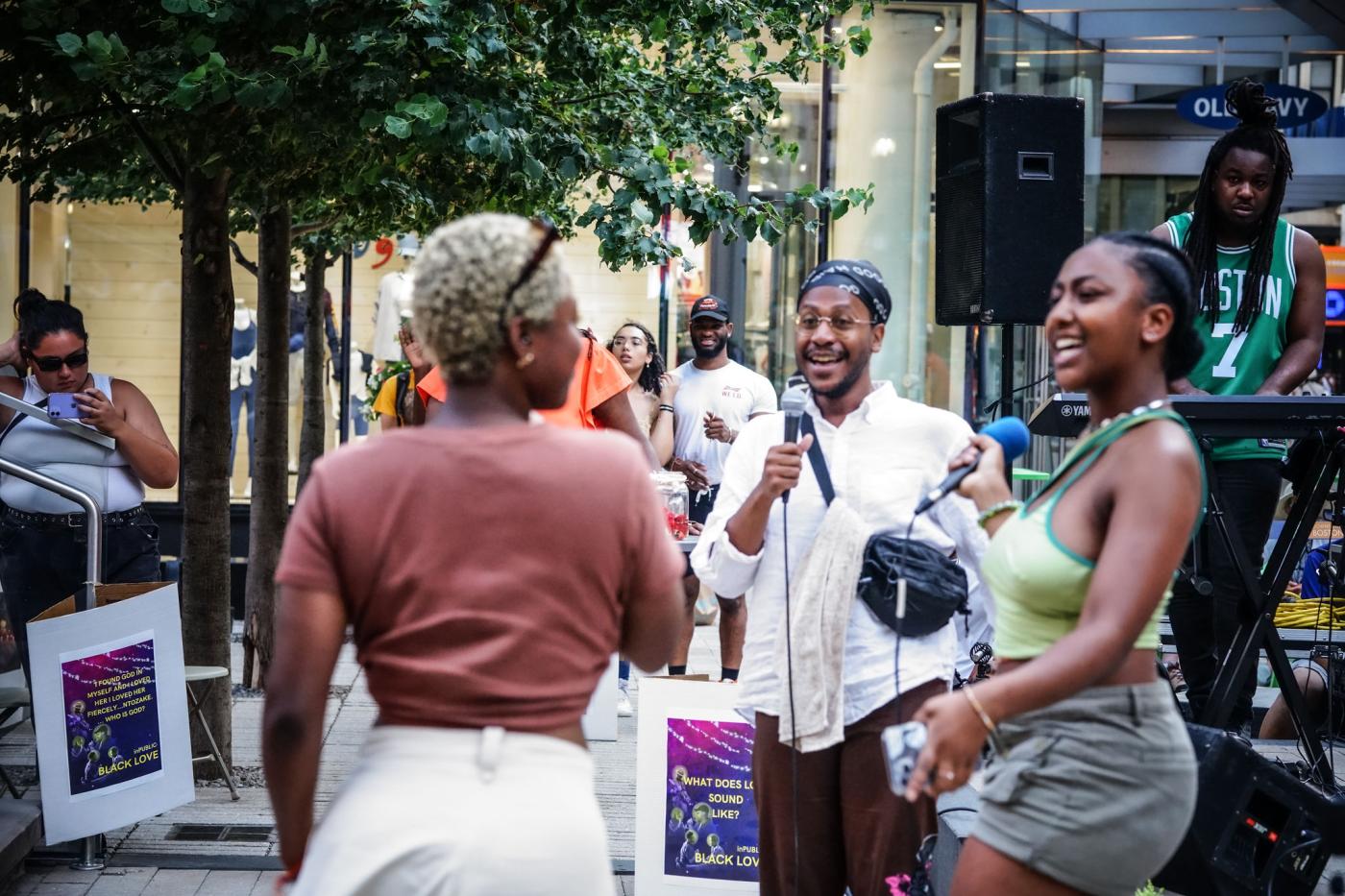 On a city street, three folks sing into microphones and look at one another.