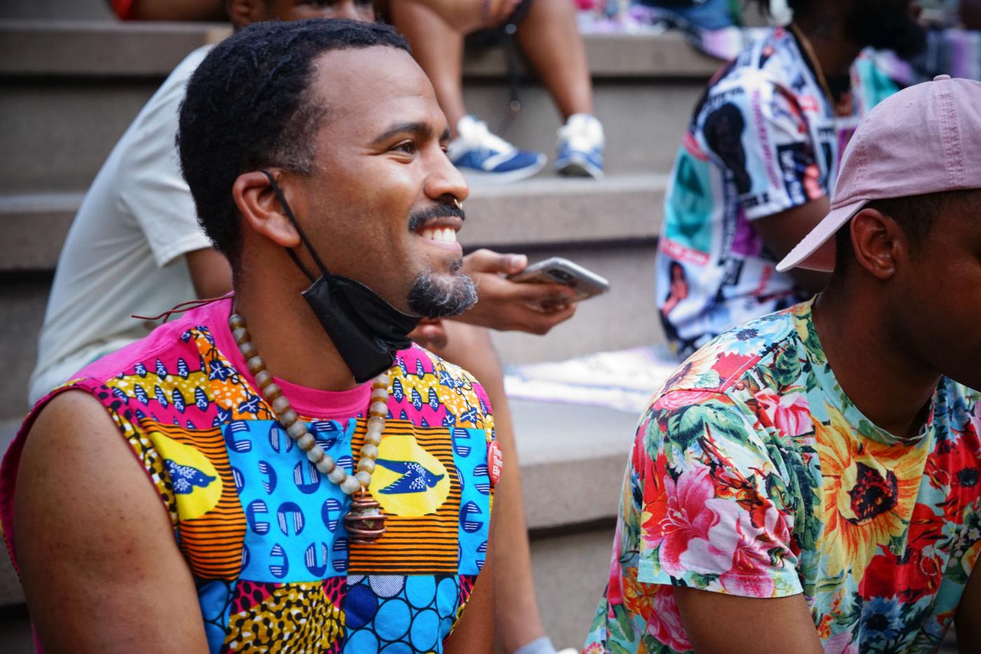A Black man smiles, has a facemask around his chin, and wears a tank top with a brightly colored print of birds and circles.