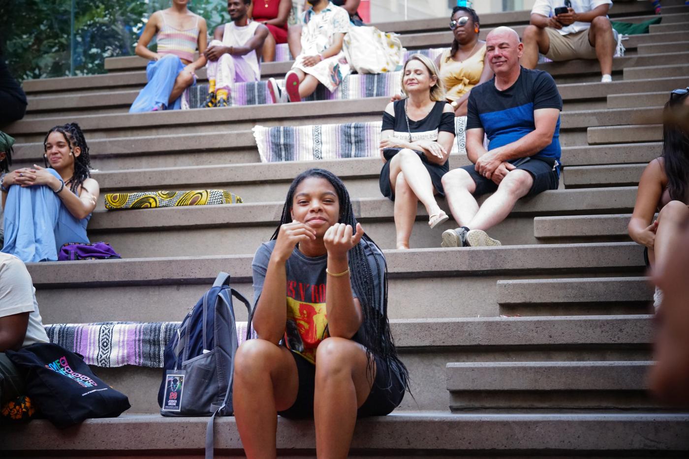 A young Black girl smiles. Behind her, a crowd of folks watch something while all sitting on stone steps.