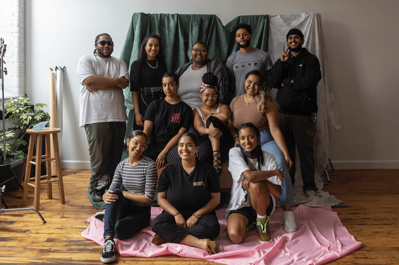 Twelve folks of color pose by some fabric laid o the ground and behind them.