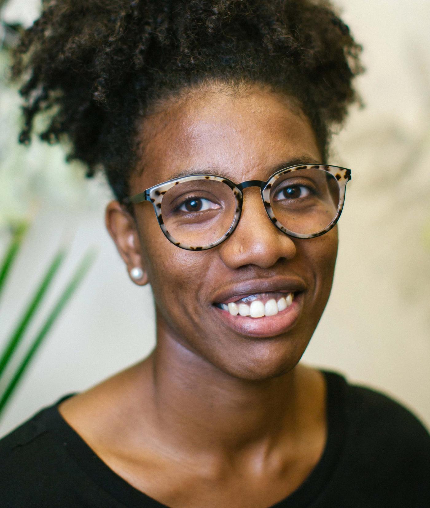A black woman with round glasses wearing a black top with her hair pulled back; ferns appear in the background.