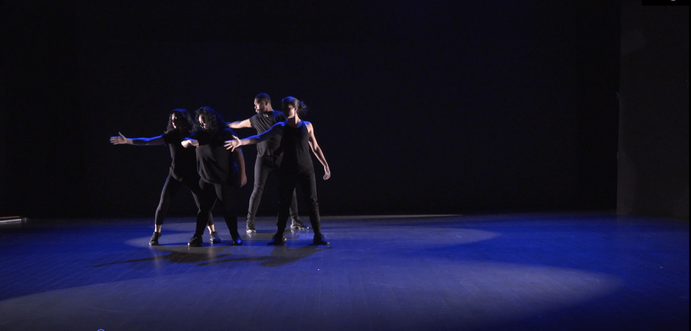 On a dim stage, four dancers, in all black, hold their arms out.