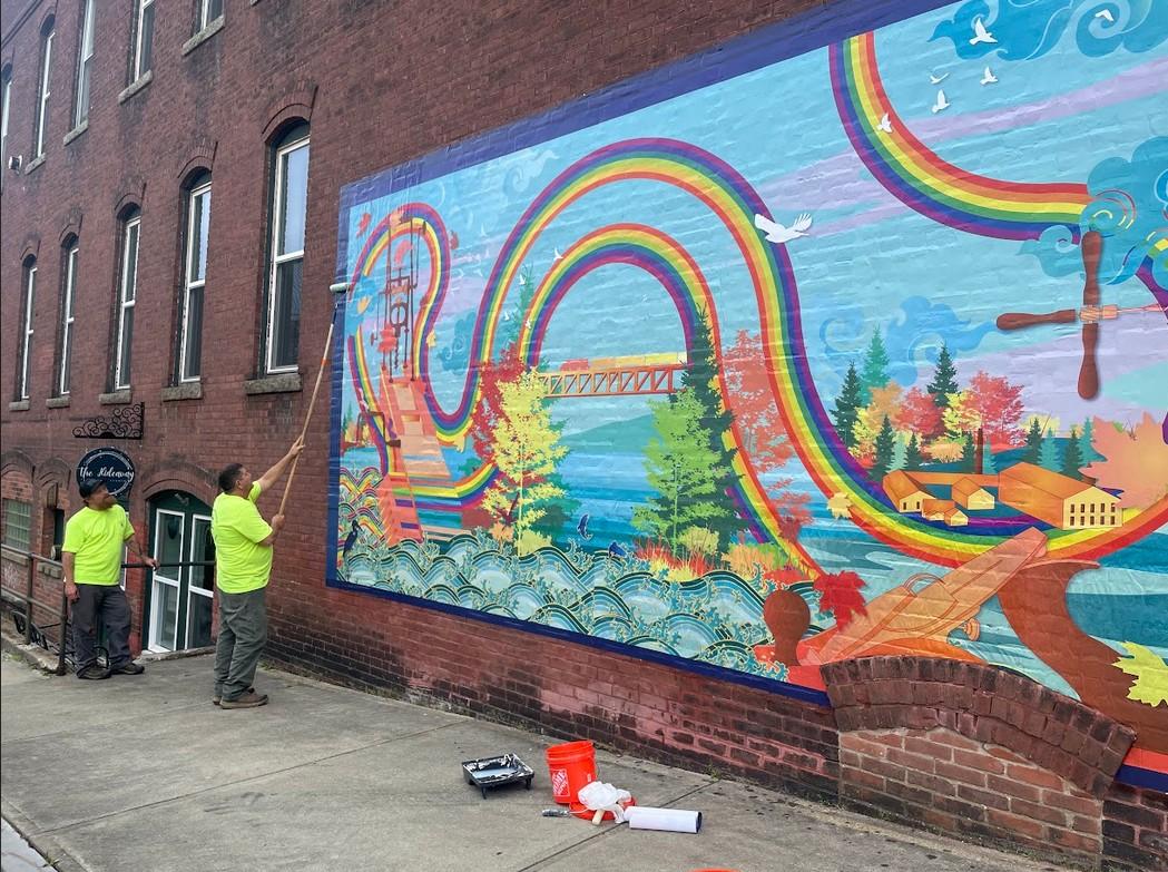 Two folks in neon shirts apply a mural to the side of a brick building. The mural is a fantasy world of rainbows that look like slides connecting rural and suburban communities. 