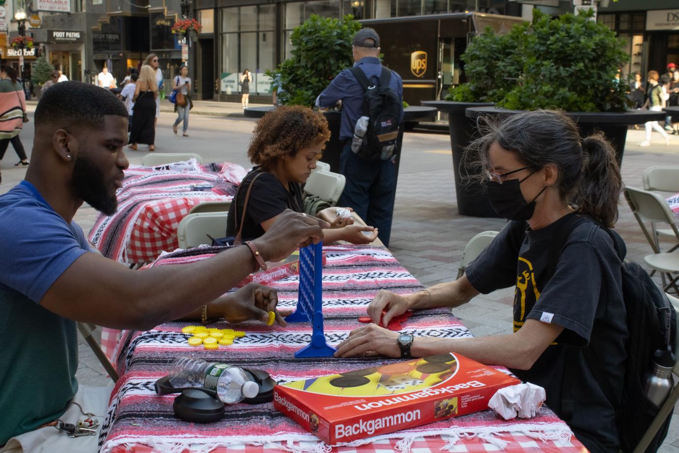 At a table, a white woman, in a face mask, and a Black man, in a blue t-shirt, play a game.