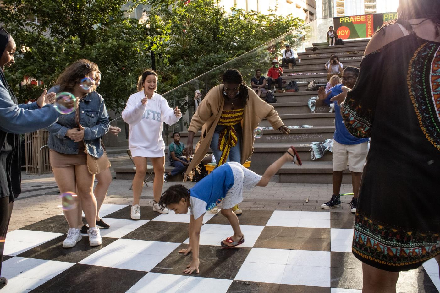 In a circle of folks, a young Black boy does a hand stand on top of a black and white checker board ground.
